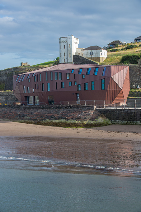 Moderne, geformte Architektur am Wasser mit einem wallenden Gebäude in Rostrot, im Hintergrund eine weiße, historische Festung und grüne Hügel.
