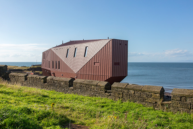 Moderne rotbraune Architektur am Meer, umgeben von Gras und alten Steinen.