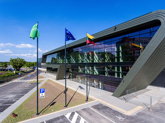 Moderne Architektur eines Gebäudes mit großen Fensterflächen und mehreren Fahnen, darunter die Europaflagge und die litauische Flagge. Das Gebäude befindet sich in einer städtischen Umgebung mit einem Parkplatz.