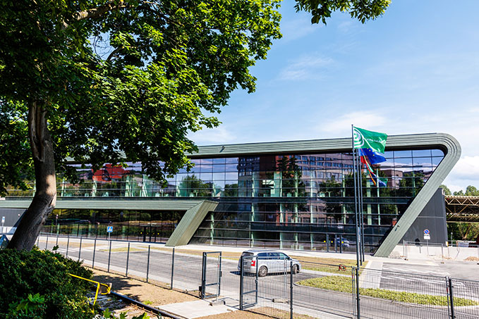 Moderne Gebäudefassade mit großen Glasfenstern und einzigartiger Architektur, umgeben von Bäumen und einem Parkplatz.