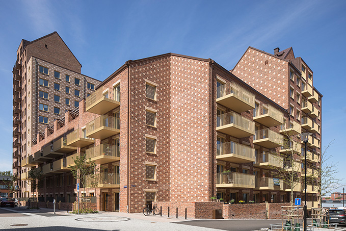 Moderne Wohnanlage mit roten Ziegeln und Balkonverkleidungen, umgeben von Bäumen und einem klaren blauen Himmel.