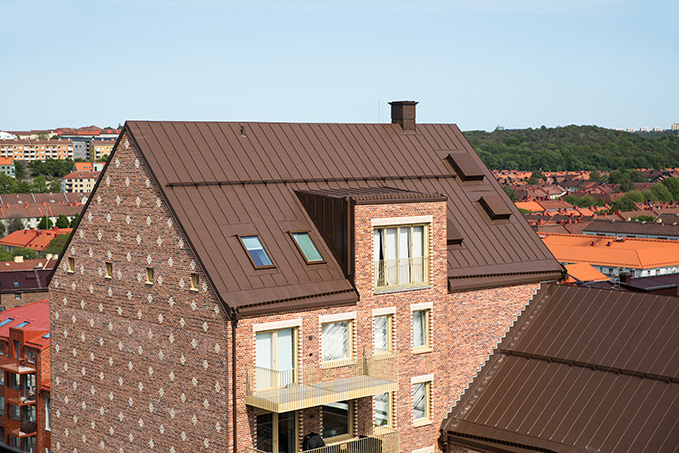 Dachansicht eines modernen Hauses mit braunem Metalldach und Ziegelfassade, umgeben von weiteren Gebäuden und grünen Hügeln im Hintergrund.